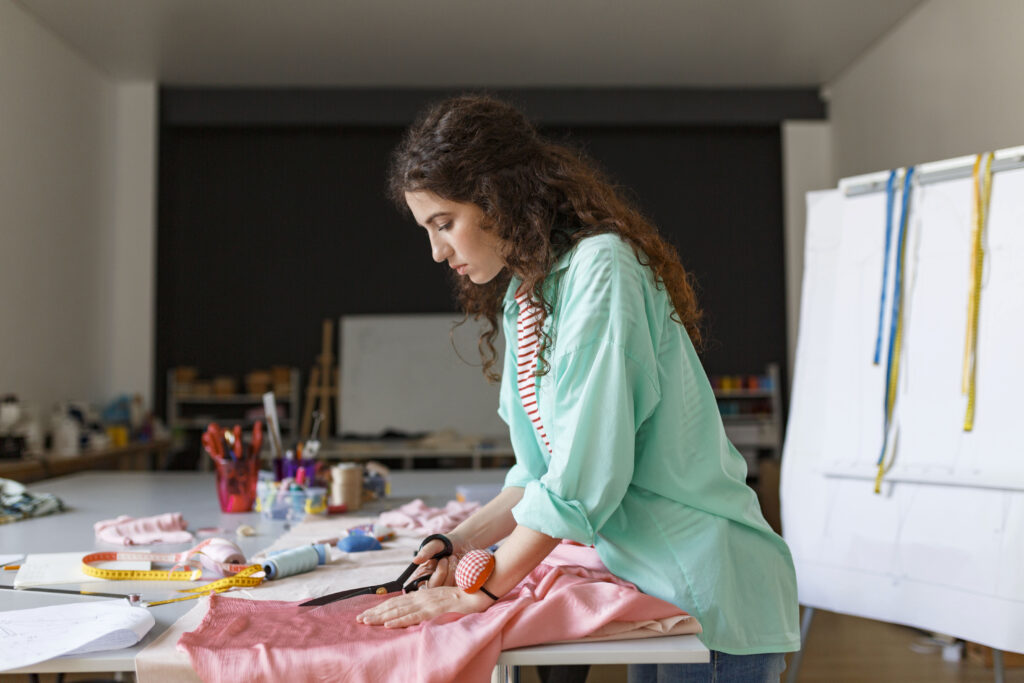 Mulher costurando em uma máquina de costura, rodeada por tecidos reaproveitados e moldes.