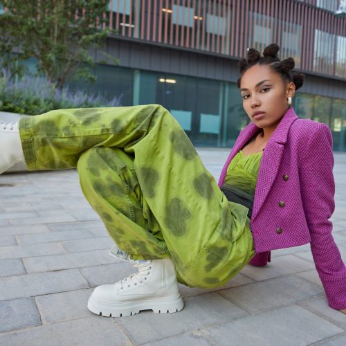 Horizontal shot of stylish teenage girl wears fashionable cloths and white boots has bright makeup poses against urban building poses for making photo practices breakdancing looks seriously.
