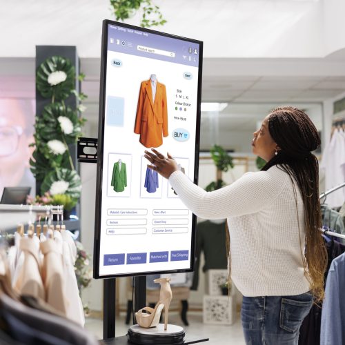 Woman buyer selecting clothes on board, using interactive touch screen monitor in clothing store. Young boutique customer buying fashion items from self ordering kiosk service, retail shop.