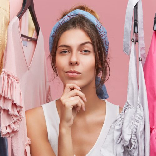 Portrait of thoughtful woman with scarf on head, standing near hangers with clothes, thinking over what to buy. Female shopaholic in boutique with pensive expression, having difficult choice
